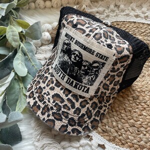 May include: A trucker hat with a leopard print pattern in shades of brown and tan. The hat features a black mesh back and a patch with the text "Mount Rushmore State South Dakota" and an image of the Mount Rushmore presidents.