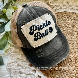 May include: A black distressed trucker hat with a beige mesh back. The hat has a white patch with the text "Pickle Ball" in black cursive lettering and a black pickleball graphic.