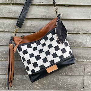 Puede incluir: Bolso de hombro con estampado de cuadros blancos y negros y detalles en cuero marrón. El bolso tiene una base de cuero negro y una parte superior de cuero marrón. Una correa negra y un adorno de pluma están adjuntos.