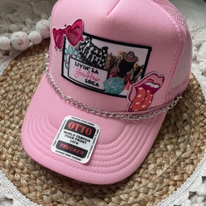 May include: Pink trucker hat with a silver chain, featuring a variety of colorful patches including a pair of sunglasses, a disco ball, a tongue, and a patch that says "Livin' La Yus Haw Loca".