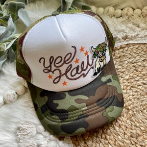 May include: A camouflage trucker hat with a white front panel featuring the text "Yee Haw" in brown rope-style lettering, orange stars, and a floral-crowned Highland cow graphic. The hat has a green mesh back and a curved brim.