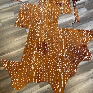 May include: A large, irregularly shaped piece of animal hide featuring a vibrant reddish-brown base color covered in a dense pattern of white spots. The hide is laid out flat, displaying its natural, organic edges and textured surface throughout.
