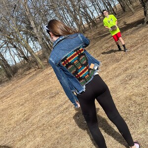 May include: A denim jacket with a colorful patterned back panel. The jacket has a classic design with a frayed hem. The back panel features a geometric design in shades of red, orange, green, and blue on a black background. The person is wearing black leggings.