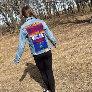 May include: A blue denim jacket with a colorful geometric pattern on the back. The pattern is reminiscent of a Native American design.