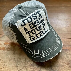 May include: A distressed olive green baseball cap with a white mesh back. The cap features a white patch with the text "JUST A SMALL TOWN GIRL" in black lettering. The brim has white stitching.