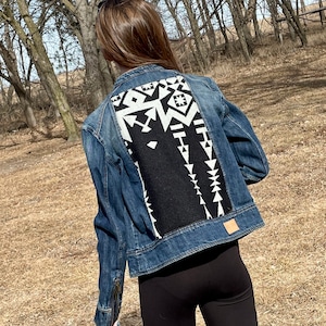 May include: A denim jacket with a black and white geometric design on the back. The jacket is a medium wash blue with a classic collar, button closure, and side pockets. The design features a mix of shapes.