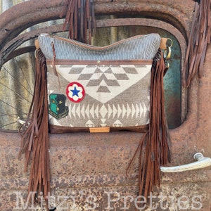May include: A brown leather fringe bag with a white and gray geometric pattern. The bag has two patches, one with a star and the other with a military insignia. The bag is sitting on a rusty metal surface.