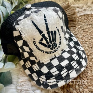 May include: Black and white checkered trucker hat with a patch that says "Expensive & Talks Back, Difficult & Talks Back" and a skeleton hand making the peace sign.