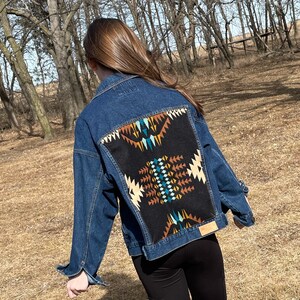 May include: A dark wash denim jacket with a large, colorful geometric design on the back. The design features a black background with tan, teal, and brown patterns. The jacket has a classic collar and button closures.