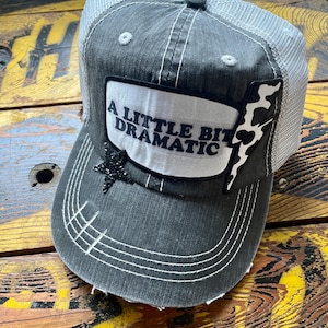 May include: A gray trucker hat with a white mesh back. The front features a white patch with the text "A LITTLE BIT DRAMATIC", a black star, and a cow print design. White stitching accents the brim.