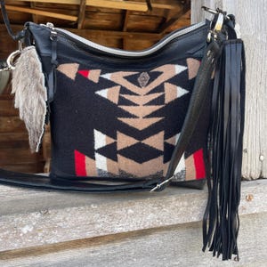 Puede incluir: Bolso bandolera de cuero negro con un estampado geométrico en beige, blanco y rojo. Cuenta con una cremallera plateada, una larga correa de cuero negro y una borla decorativa. Un adorno de piel cuelga de un lado.