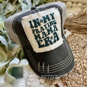 May include: A distressed black trucker hat with a gray mesh back. The front patch features the text "IN MY PRAYING MAMA ERA" in teal lettering. The hat has a curved brim and white stitching details. The brand name "MITZI'S PRETTIES" is printed on the brim.