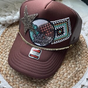 May include: A brown trucker hat with a mesh back and a curved brim. The hat features a sparkly star, a circular patch with text, and a rectangular patch with a geometric design. A gold chain with small discs accents the brim.