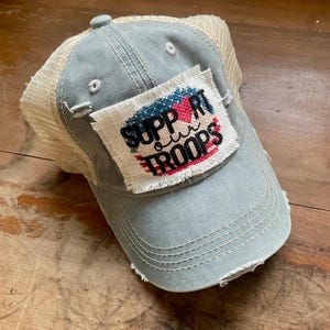 May include: A distressed, light blue baseball cap with a beige mesh back. The cap features a patch with the words "SUPPORT our TROOPS" in a patriotic design with red, white, and blue colors. The brim is slightly frayed.