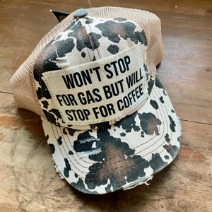 May include: A trucker hat with a cow print design in brown, black, and white. The hat features a mesh back and a distressed patch with the text "WON'T STOP FOR GAS BUT WILL STOP FOR COFFEE".