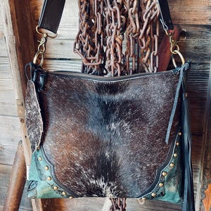 Puede incluir: Bolso bandolera de cuero de vaca marrón y negro con correa negra y herrajes dorados. El bolso presenta un detalle turquesa con tachuelas doradas y flecos negros. Cierre con cremallera.