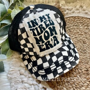 May include: Black and white checkered trucker hat with a black patch that says "In My Tired Mom Era" in a retro font.