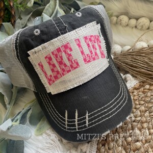 May include: Distressed charcoal gray trucker hat with a white mesh back. The front features a white patch with the words "LAKE LIFE" in pink cross-stitch. The hat has a curved brim and white stitching details.