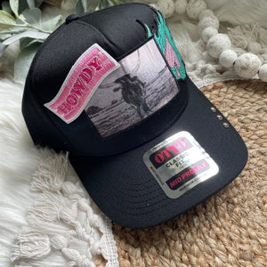 May include: Black baseball cap with a pink "Howdy" patch, a black and white photo patch of a cowboy roping a steer, and a green and blue flame patch. The cap has a black brim with a silver "OTTO" label that says "Classic Fit" and "Mid Profile".
