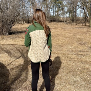 May include: Green jacket with a floral patterned back panel. The jacket has a patch on the left sleeve and a brown leather accent. The person is wearing black pants.