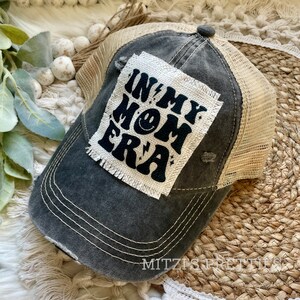 May include: Distressed gray baseball cap with a tan mesh back. The cap features a patch with the text "IN MY MOM ERA" in black, along with a smiley face and lightning bolt graphics. The hat has a pre-worn look with visible stitching.