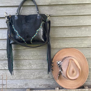 Puede incluir: Bolso de cuero negro con detalles en turquesa y dorado y flecos. El bolso está colgado en una pared de madera junto a un sombrero de vaquero de cuero marrón con una banda de piel de serpiente.
