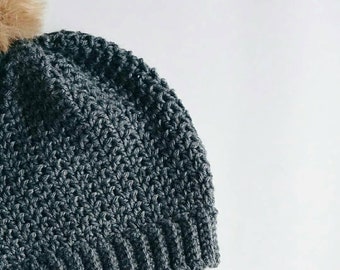 May include: A dark gray crocheted beanie with a brown faux fur pom pom on top.