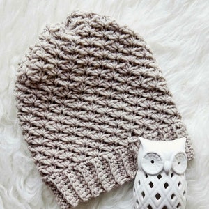 May include: A beige crocheted beanie hat with a textured stitch pattern. The hat is sitting on a white fluffy surface next to a white ceramic owl figurine.