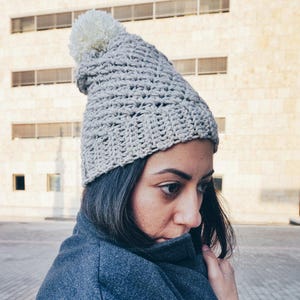May include: A gray crocheted beanie with a white pom pom. The beanie is worn by a person with long dark hair and a gray coat.