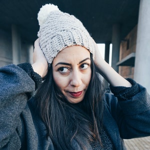 May include: A woman wearing a gray knitted beanie with a white pom pom, a gray coat, and a black and white patterned shirt.