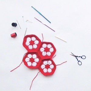 May include: A collection of crochet supplies on a white surface. The image features red and white crocheted hexagons, crochet hooks, needles, pins, scissors, and spools of thread. The hexagons are arranged in a cluster, showcasing the craft's vibrant colors.