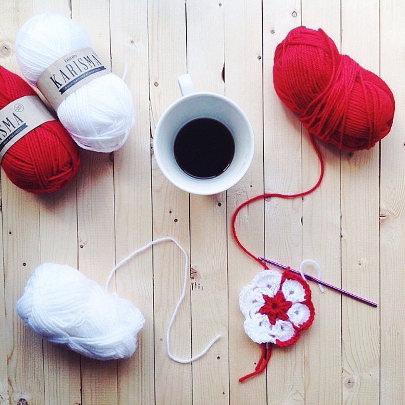 May include: Three skeins of Drops Karisma yarn in solid white and red, a white ceramic mug filled with dark liquid, and a partially crocheted flower motif in red and white yarn with a metal crochet hook resting on a light wood surface.