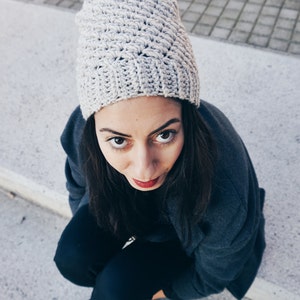 May include: A woman wearing a gray crocheted beanie with a white pom pom, a dark gray jacket, and black pants. She is sitting on a concrete surface and looking at the camera.
