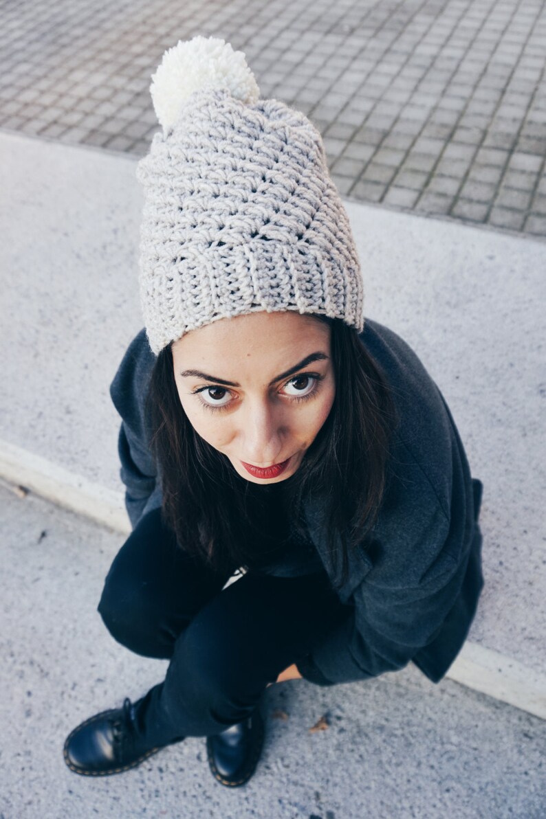 May include: A woman wearing a gray crocheted beanie with a white pom pom, a dark gray jacket, and black pants. She is sitting on a concrete surface and looking at the camera.