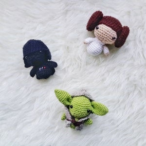 May include: Three handmade crochet dolls: a black figure with a red and white design, a light-skinned figure in a white dress with brown hair buns, and a green figure with large ears and a gray cloak. The dolls are on a white, fluffy surface.
