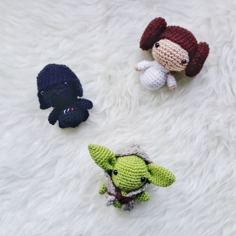 May include: Three handmade crochet dolls: a black figure with a red and white design, a light-skinned figure in a white dress with brown hair buns, and a green figure with large ears and a gray cloak. The dolls are on a white, fluffy surface.