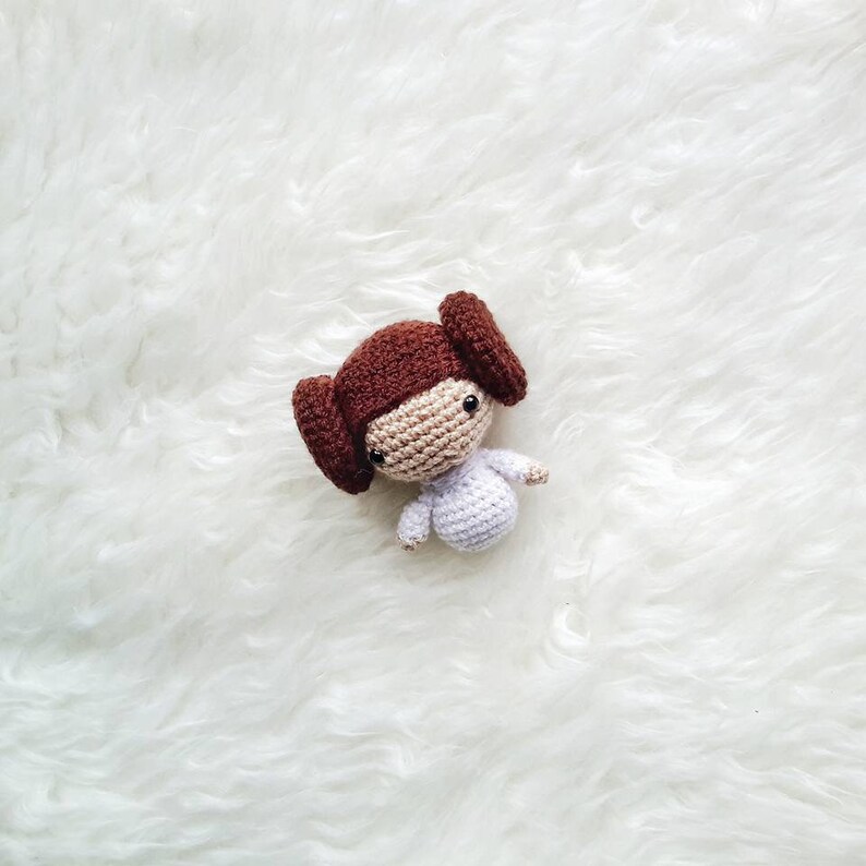 May include: A handmade crochet doll of a character with brown hair styled in two side buns. The doll has a tan face, black eyes, and is wearing a white dress. The doll is approximately 4 inches tall.