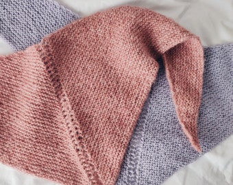 May include: Two knitted triangular scarves, one in a light pink color and the other in a light purple color, are folded together on a white surface.
