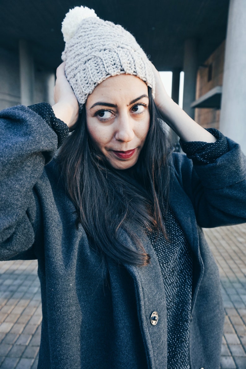 May include: A woman wearing a gray knitted beanie with a white pom pom, a gray coat, and a black and white patterned shirt.