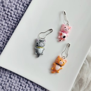 Op de afbeelding: Drie kleurrijke kattenvormige steekmarkeerders met veiligheidsspelden. De katten zijn grijs, roze met zwarte vlekken en oranje met zwarte strepen.
