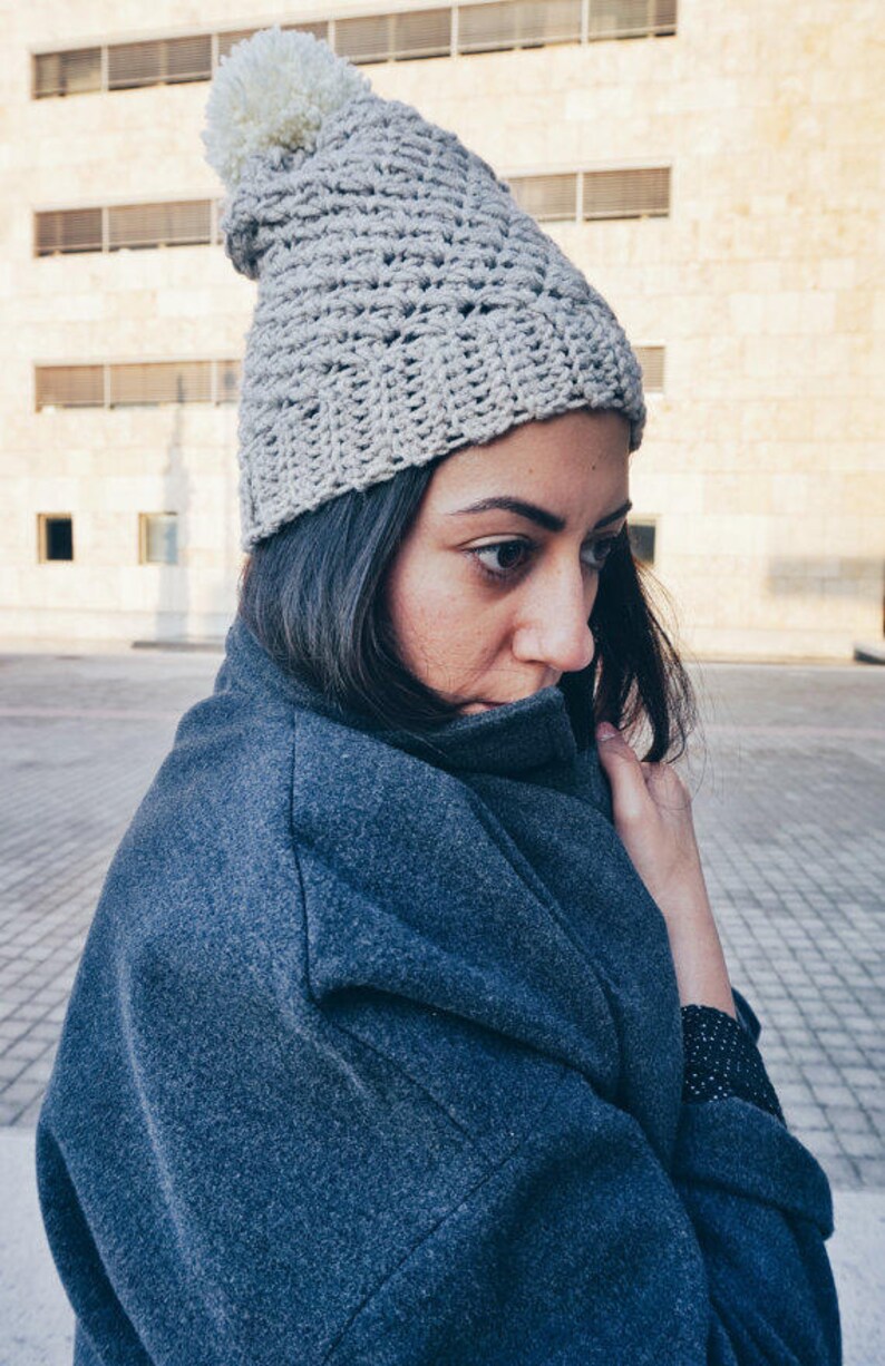 May include: A gray crocheted beanie with a white pom pom. The beanie is worn by a person with long dark hair and a gray coat.