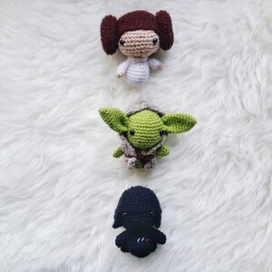 May include: Three handmade crochet dolls: a brown-haired princess, a green-skinned Jedi Master, and a black-clad Sith Lord. The dolls are crafted with detailed features and are arranged vertically on a white, fluffy surface. The dolls are approximately 3-4 inches tall.