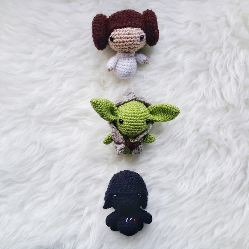 May include: Three handmade crochet dolls: a brown-haired princess, a green-skinned Jedi Master, and a black-clad Sith Lord. The dolls are crafted with detailed features and are arranged vertically on a white, fluffy surface. The dolls are approximately 3-4 inches tall.
