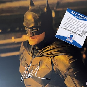 Puede incluir: Una fotografía de Batman con un traje negro y una firma blanca en la parte inferior. Una tarjeta de certificado de autenticidad de Beckett está colocada en la esquina superior derecha, con el texto Beckett Certificate of Authenticity y BD57531.