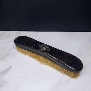 Könnte beinhalten: Eine rechteckige Kleiderbürste mit einem dunkel schwarzen Holzgriff und sichtbarer Holzmaserung. In der Mitte des Griffs befindet sich ein kleines, verziertes silberfarbenes Metallemblem. Die Bürste hat dichte, hellbraune Naturborsten.