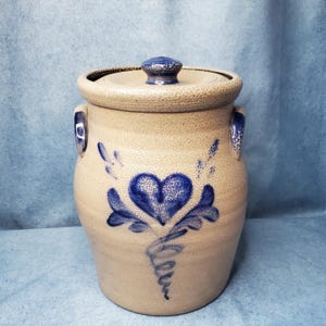 May include: A tan stoneware crock with a lid, featuring a blue hand-painted heart design on the front. The cylindrical container has two side handles and a textured, speckled surface finish. The lid includes a small, rounded blue knob handle.