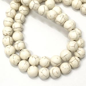 Puede incluir: Una hebra de cuentas de howlite blancas con forma redonda y lisa, con un patrón natural veteado. Las cuentas están enhebradas en un hilo transparente.