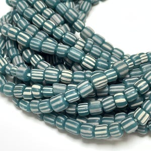 May include: A close-up of a strand of teal blue and white striped glass beads. The beads are small and cylindrical, with a glossy finish.