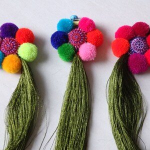 Pode incluir: Três broches de flores de pompom coloridas com borlas verdes. Cada broche apresenta um círculo central com bordado e é feito de uma variedade de cores, incluindo rosa, roxo, azul, verde, laranja e vermelho.