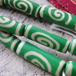 May include: Close-up of green and cream-colored wooden beads with a spiral design. The beads are strung together with white string. The background is a light-colored, textured surface.
